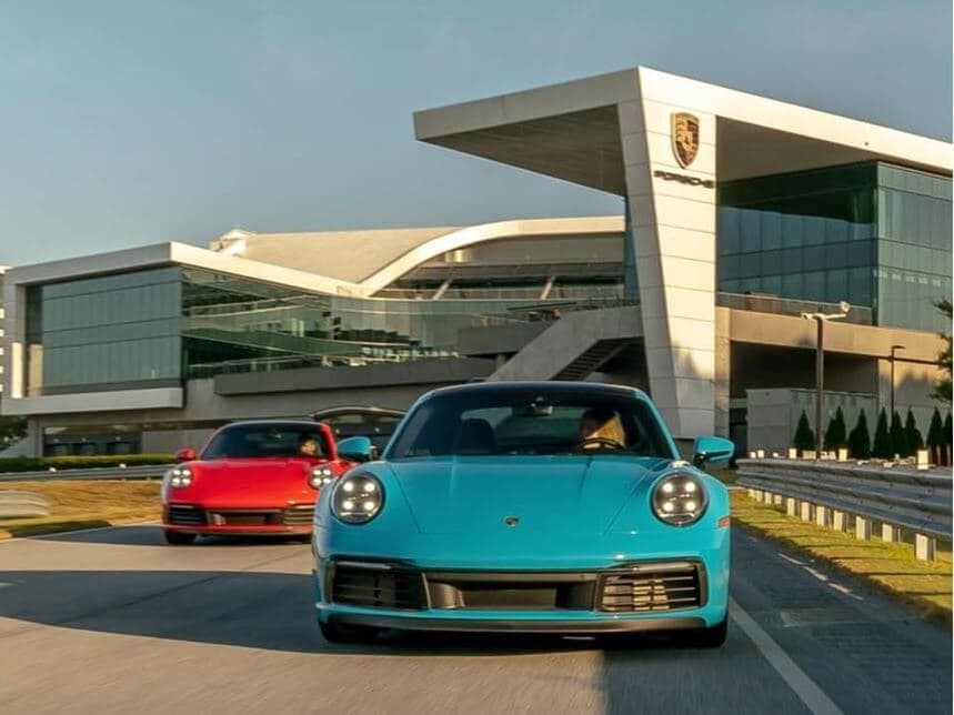 Two sports cars, one blue and one red, drive in front of Porsche Experience Center Atlanta on a sunny day.
